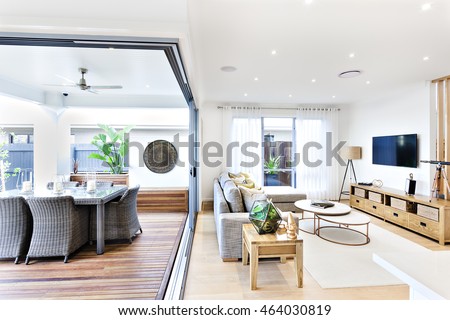 Modern living room attached to the patio and dining area including a plastic chairs and tables on the wooden floor outside near to the sofa and pillows on the carpet beside the television and cabinet