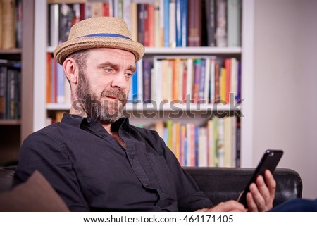 Hipster with hat sitting on a couch in his liviing room with a mobile phone