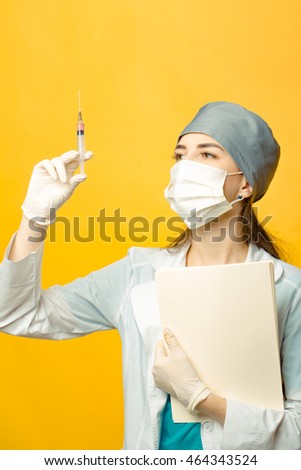 Beautiful young doctor in mask and medical uniform holding syringe with liquid and folder on yellow background