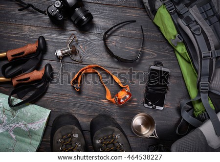 Hiking accessories set on dark wooden background:  boots, backpack, sunglasses, photo camera, map, smartphone, flashlight and others. Top view