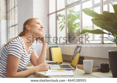 Young woman sitting daydreaming at the office leaning her chin on her hand and staring up out of a nearby window with a dreamy smile of pleasure Royalty-Free Stock Photo #464538905
