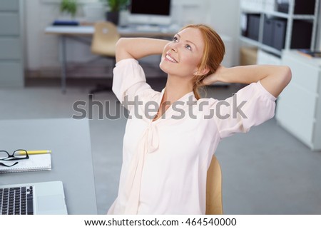 Successful businesswoman sitting daydreaming at her desk in the office with her hands clasped behind her head looking up with a happy smile