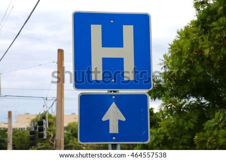 Blue and White Hospital Sign H Symbol Straight Ahead Arrow Pointing Up with Cloudy Sky, Power Lines, Buildings and Trees in the Background