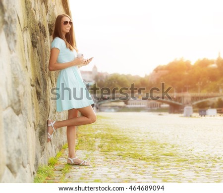 Beautiful young brunette woman listening music in headphones, holding a cup of coffee and enjoying music. Old town center with river on background