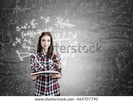 Young girl with long hair holding book and smiling standing near blackboard with formulas on it. Concept of exact sciences studying 