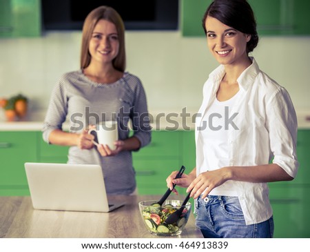 Beautiful girl is mixing salad, looking at camera and smiling while cooking in kitchen at home. Her friend is holding a cup in the background