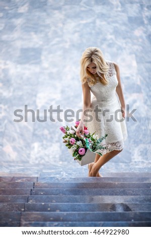 Young beautiful blond woman in a white dress holding a basket of flowers and stands on a marble staircase.