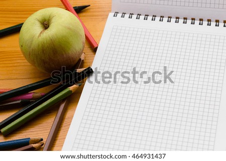 Variegated colored pencils, white square-lined paper notebook and green apple on a light orange wooden study table close-up