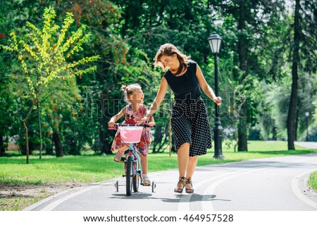 Beautiful and happy young mother teaching her daughter to ride a bicycle. Both smiling and looking at each other. Summer park in background. Royalty-Free Stock Photo #464957528