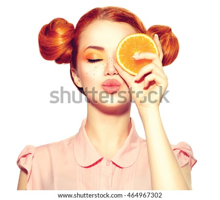 Beauty Model Girl holding Juicy Orange slice. Beautiful Joyful teen girl with freckles, funny red hairstyle and yellow makeup. Professional make up. Isolated on white background