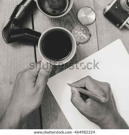 Black and white top view close-up picture of human hands working writing, wooden table background. The following objects are used on the table for work: map, paper, film camera, coffee, compass, etc.