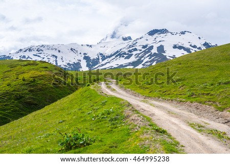 Dirt road in a highland. Road goes through summer pastures full of green grass. The foot of Ushba mountain. Snow-capped mountains in the background