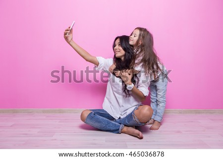 beautiful young mother and her cute daughter in white shirts and jeans sit on the floor on a pink background wall and make selfie Royalty-Free Stock Photo #465036878