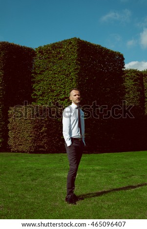 Male fashion concept. Handsome mature man with silver beard posing over green bushes of city park. Hands in pockets. Warsaw, Poland, Arkady Kubickiego. Graphic, geometric composition. Minimalism style