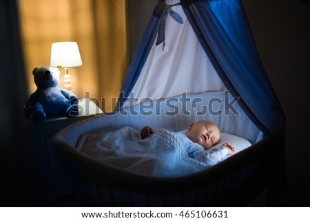 Adorable baby sleeping in blue bassinet with canopy at night. Little boy in pajamas taking a nap in dark room with crib, lamp and toy bear. Bed time for kids. Bedroom and nursery interior. Royalty-Free Stock Photo #465106631