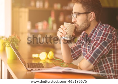 Toned picture of handsome freelance man in glasses drinking delicious coffee while working in restaurant or cafe on laptop computer. Business or freelance concept.
