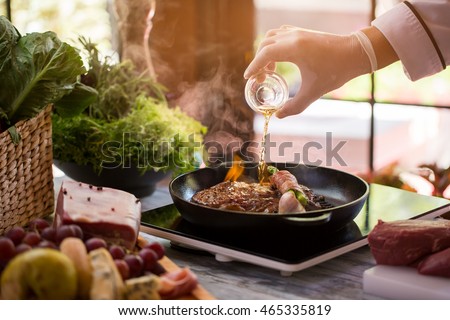 Bottle pours liquid onto pan. Meat and green vegetable. Fried beef and asparagus. Chef makes flambe steak. Royalty-Free Stock Photo #465335819