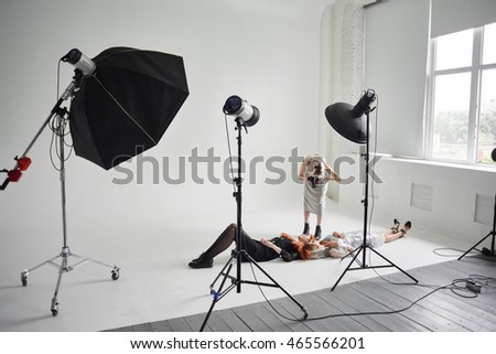 Girl photographer photographing fashion models in a clear coat with a pomegranate mask on the face and black clothes lying on white floor in Studio
