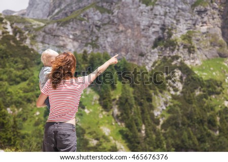 Portrait of happy young loving mother and her baby outdoors looking forward and pointing. Beautiful woman with a child on hands standing in the forest and mountains. Travel, explore, family concept