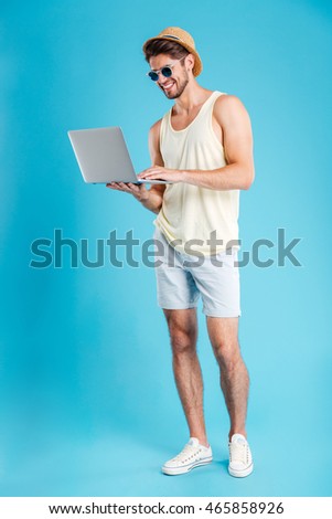 Full length of happy young man in hat and sunglasses standing and using laptop over blue background