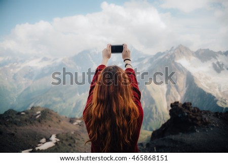 beautiful young woman with red hair photographer takes a picture of a mountain landscape on the phone while hiking