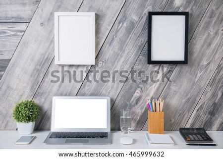 Creative designer workplace with blank white laptop computer, plant, smartphone, water glass, calculator, stationery items and two picture frames hanging above on wooden background. Mock up
