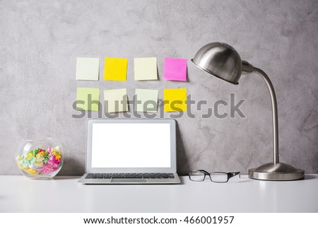 Creative hipster workplace with blank white laptop computer, decorative glass bowl, glasses, table lamp and colorful stickers on concrete wall background. Front view, Close up, Mock up