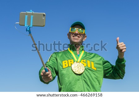 Gold medal athlete in Brazil jacket gives a smiling thumbs up as he poses for a celebration picture. Focus on the end of the selfie stick.