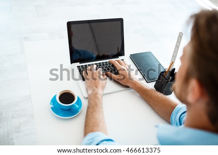 Young handsome cheerful confident pensive businessman sitting at table working on laptop with cup of coffee aside. White modern office interior background.