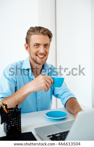 Young handsome cheerful confident businessman sitting at table working on laptop drinking coffee. Looking in camera smiling. White modern office interior background.