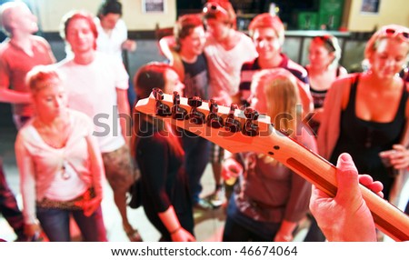 Hands on the fretboard of an electric guitar in a nightclub with people dancing in the background
