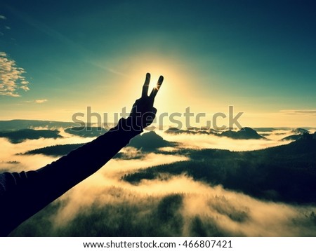 Man hand make victory to Sun. Misty daybreak in a beautiful hills. Peaks of hills are sticking out from foggy background, the fog is red and orange due to Sun rays. 