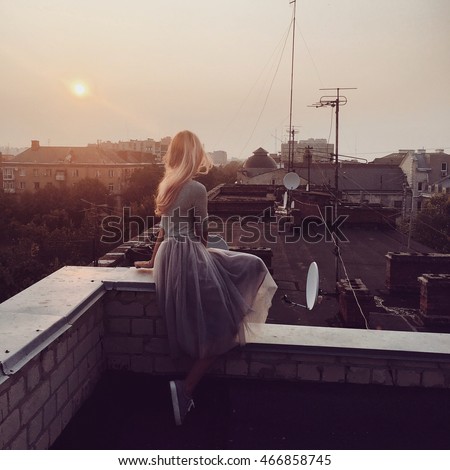 Girl in bohemian skirt is sitting on the roof. City sunset. iPhone shooting