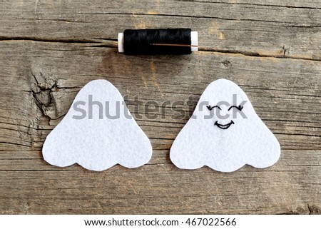 Cut from white felt details for making Halloween ghost decor, thread, needle on a wooden table. On one side embroidered with black thread eyes and mouth. A step instruction for kids. Wooden background