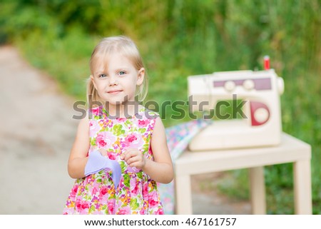 little blonde smiling lovely child 4 years old (girl) in cotton dress sewing needle (hand) of the purple cloth on a background of a sewing machine with a cotton cloth beautiful outdoors