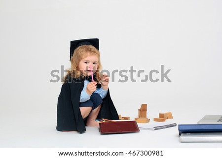 Little boy serious child in black academic gown and squared cap holding pink colored pencil and sitting near box with pencils wooden blocks diaries isolated on white background