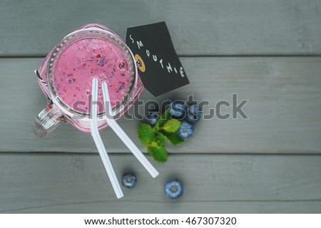 Cup of blueberry smoothie with a signed label on a table, on which lay blueberries and a sprig of mint.