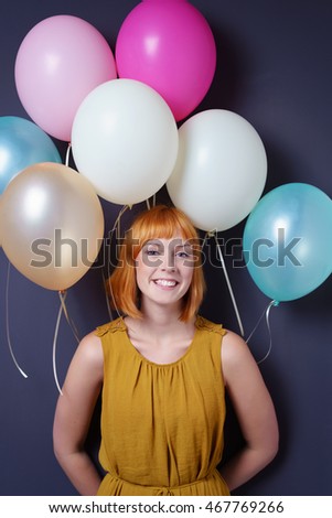 Fun young redhead woman holding a bunch of balloons behind her back standing smiling happily at the camera as she celebrates a special occasion