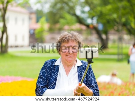 Grandmother with trendy dress and bandage holding self-ie stick to take photo in the floral garden.