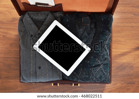 Vintage travel background. Top view of old suitcase with clothes on wooden floor, with tablet computer with copyspace. Retro style. 