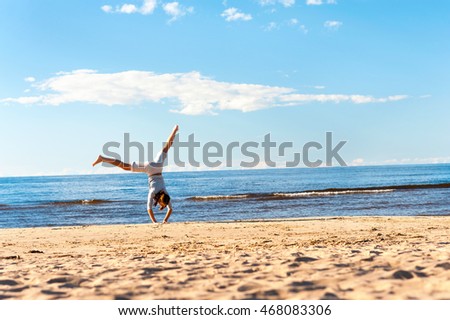 Cheerful teenage girl doing handstand on the beach sand on blue cloudy sky background. Vibrant multicolored horizontal outdoor summertime image with copy space. Latvia.
