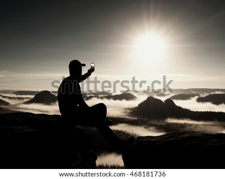 Hiker in black sportswear is sitting on cliff's edge. Man taking nature photos by smart phone while sunrise above mountains. Vivid effect