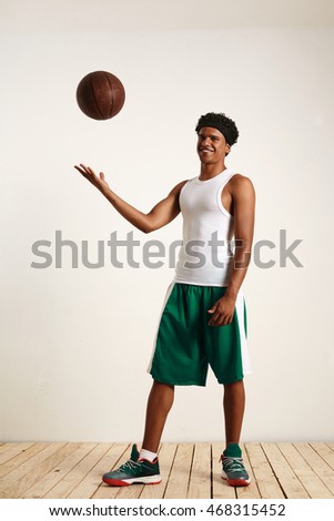 Full length portrait of a muscular black attractive male in basketball green and white outfit with a grunge brown basketball in the air against white wall and wooden floor.