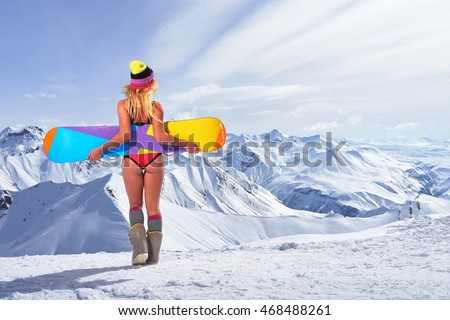 Back view of unrecognizable blonde girl in bikini swimsuit with snowboard in outstretched arms above head against of snowy mountains in sunlight