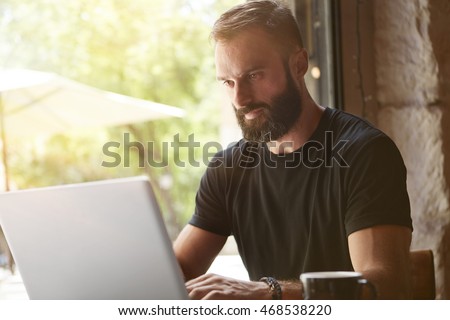 Concentrated Bearded Man Wearing Black Tshirt Working Laptop Wood Table Urban Cafe.Young Manager Work Notebook Modern Interior Design Loft Place.Coworking Process Business Startup.Blurred Background