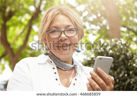 Headshot of beautiful Caucasian female pensioner looking at camera with happy cheerful expression while using cell phone for communicating online with her friends, reading news, sending photos