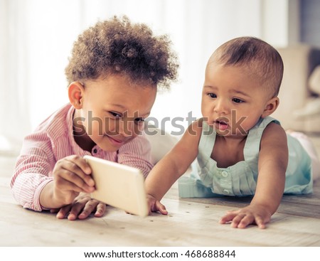 Beautiful little Afro American girls are playing on floor at home. Older girl is showing a smartphone her baby sister Royalty-Free Stock Photo #468688844