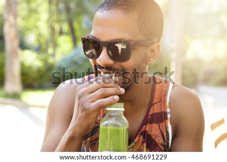 Street lifestyle concept. Young smiling African American male with moustache and short beard drinking fresh juice during date, dressed casually in colorful tank top and trendy shades or sunglasses