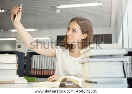 Human and technology. People and education. Indoor portrait of Caucasian teenage girl at library trying to take selfie, surrounded by books and manuals, looking cheerful. Selective focus on the woman