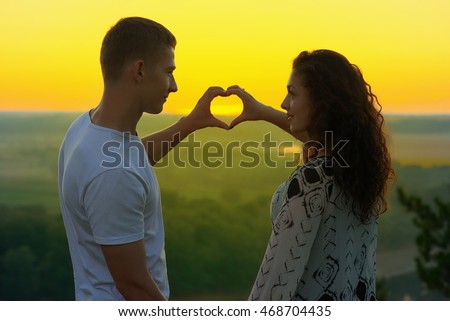 romantic couple at sunset show a heart shape from hands, beautiful landscape and bright yellow sky, love tenderness concept, young adult people